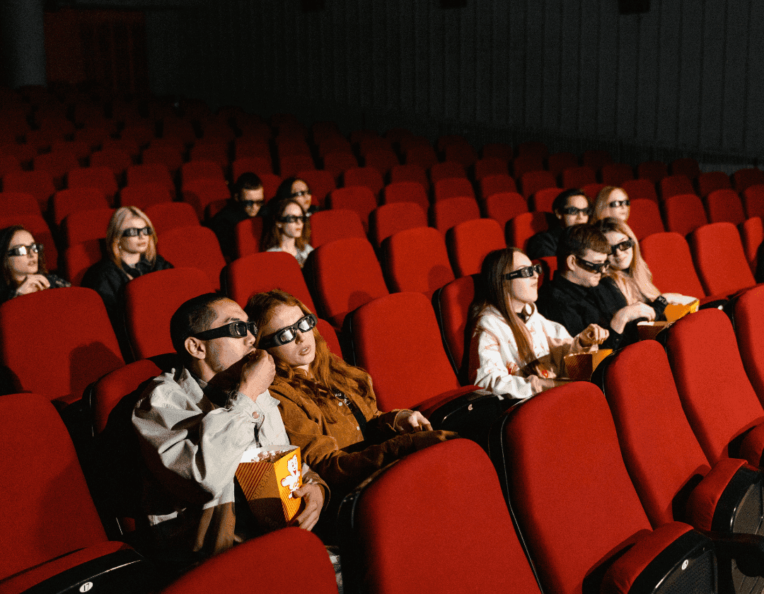 An audience in 3D glasses watching a film from red cinema seats, popcorn in hand