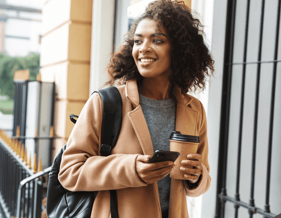 A smiling woman with a backpack and a coffee checking her phone on a city street