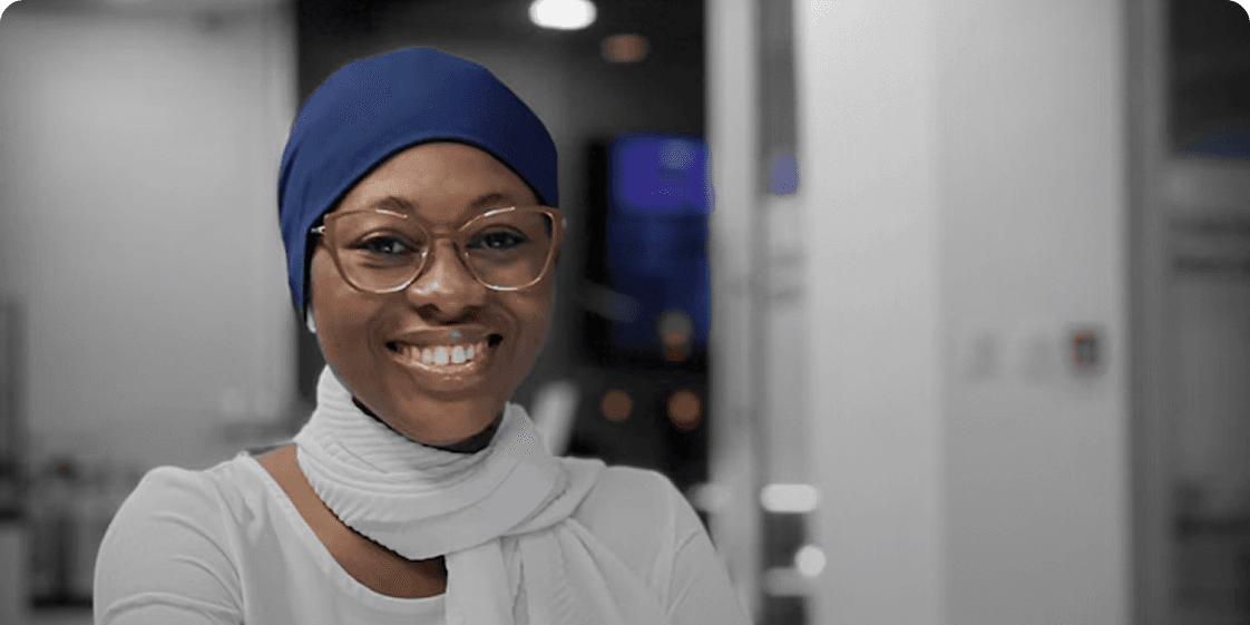 A smiling woman in a blue head wrap and glasses in a bright office